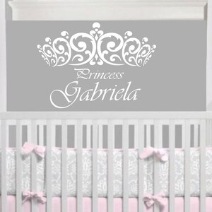 Może przedstawiać: Biała naklejka ścienna z winylu z ozdobną koroną i tekstem "Princess Gabriela" na szarym tle. Naklejka znajduje się nad białym ł&oacute;żeczkiem z r&oacute;żowo-szarym wzorzystym pościelą.