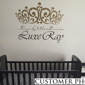 May include: A nursery wall decal featuring a gold crown design above the words "...Our Little Prince Luxe Ray" in elegant script. Below, a black crib with a black and white patterned sheet is visible.