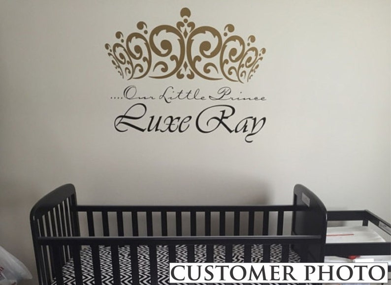 May include: A nursery wall decal featuring a gold crown design above the words "...Our Little Prince Luxe Ray" in elegant script. Below, a black crib with a black and white patterned sheet is visible.