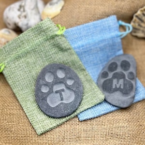 May include: Two personalized pet memorial stones with paw prints and initials T and M. The stones are in small burlap bags, one green and one blue.