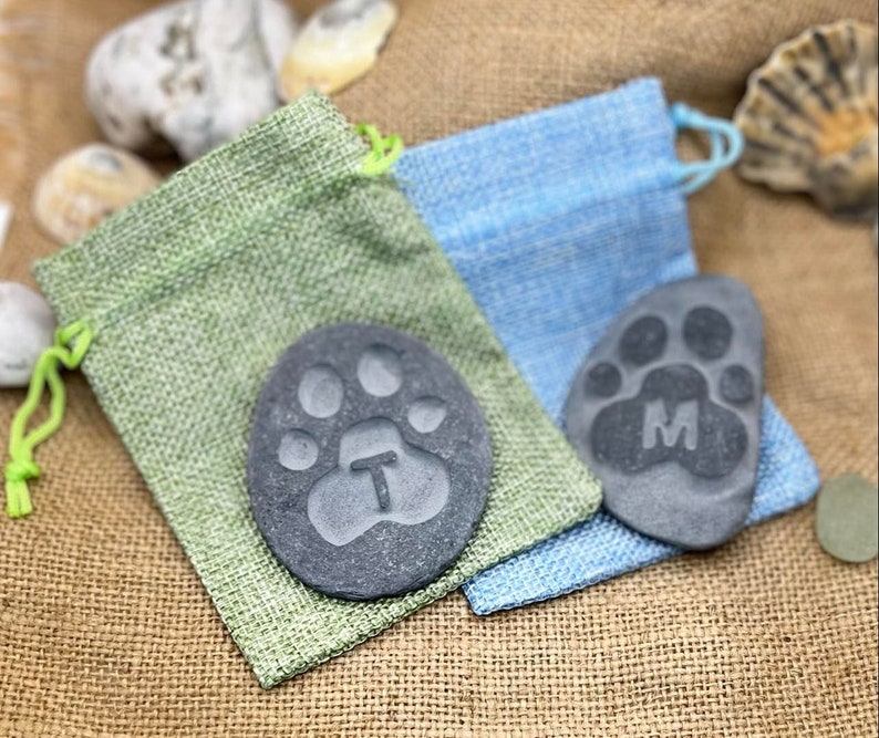 May include: Two personalized pet memorial stones with paw prints and initials T and M. The stones are in small burlap bags, one green and one blue.