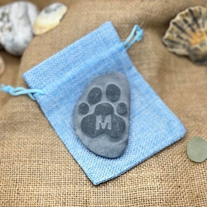 May include: A gray stone with a paw print and the letter M carved into it. The stone is in a light blue drawstring bag.
