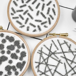 May include: Three embroidery hoops with white fabric and black embroidery designs. The designs are geometric patterns.