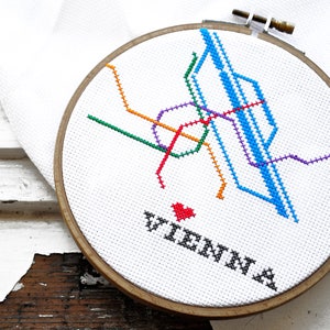 May include: Cross-stitch embroidery of the Vienna subway map in a wooden hoop. The map features lines in green, orange, purple, red, and blue. The word "VIENNA" is stitched below a red heart.