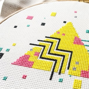 Op de afbeelding: Een geometrisch kruissteekpatroon in geel, roze en zwart in een ronde borduurring. Het patroon is een gestileerde driehoek met zigzaglijnen en een roze en gele rand.