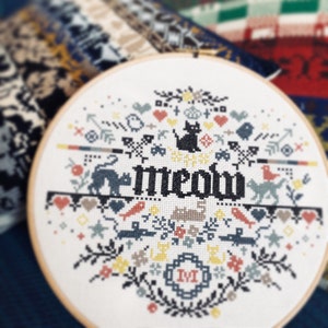 May include: A cross-stitch embroidery hoop with a cat-themed design. The word "meow" is stitched in black, surrounded by cats, birds, hearts, and floral elements in various colors. The design is set in a wooden hoop, a popular craft item.