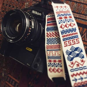 May include: A black Nikon camera with a Carl Zeiss Jena DDR lens attached. The camera has a white and red patterned camera strap with blue and red geometric designs.