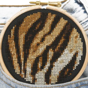May include: A close-up of a round, wooden embroidery hoop with a black fabric background. The fabric is stitched with a brown and white zebra print pattern.