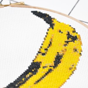 May include: A close-up of a yellow and black cross-stitch embroidery of a banana on white Aida cloth.