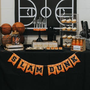 May include: A basketball-themed party setup with a black tablecloth, featuring a "SLAM DUNK" banner. The table displays basketballs, cupcakes, drinks, and treats. A basketball court graphic is in the background.
