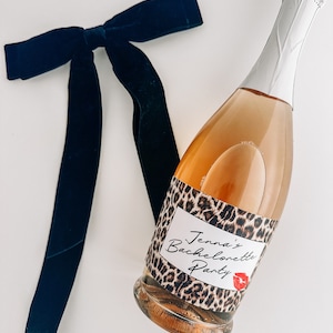 Puede incluir: Una botella de vino espumoso con estampado de leopardo y una etiqueta blanca que dice "Jenna's Bachelorette Party". Un lazo de terciopelo negro est&aacute; en el fondo.