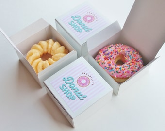 May include: Three white boxes with pink and blue striped lids. Each box has a donut shop logo with a pink and blue donut. One box contains a pink donut with colorful sprinkles, and another box contains a yellow donut with a swirl design.