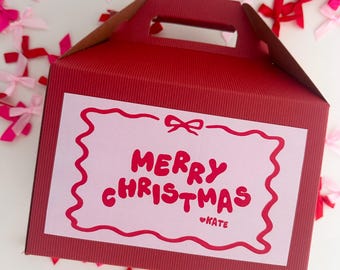 May include: A red gift box with a handle and the words "MERRY CHRISTMAS" in red on a pink label. The label has a wavy red border and the name "KATE" below the text. Scattered around the box are pink and red bows.
