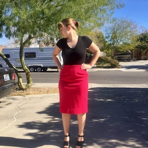 May include: A woman wearing a black v-neck top and a bright red pencil skirt. She is standing on a paved driveway with a trailer in the background.