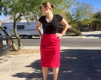 May include: A woman wearing a black v-neck top and a bright red pencil skirt. She is standing on a paved driveway with a trailer in the background.