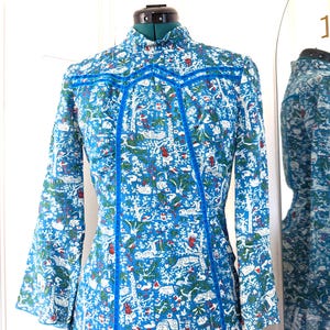 May include: A long-sleeved dress with a high collar, displayed on a mannequin. The dress features a vibrant blue background with a white, green, and red pattern of trees and animals. Bright blue trim accents the seams and neckline.