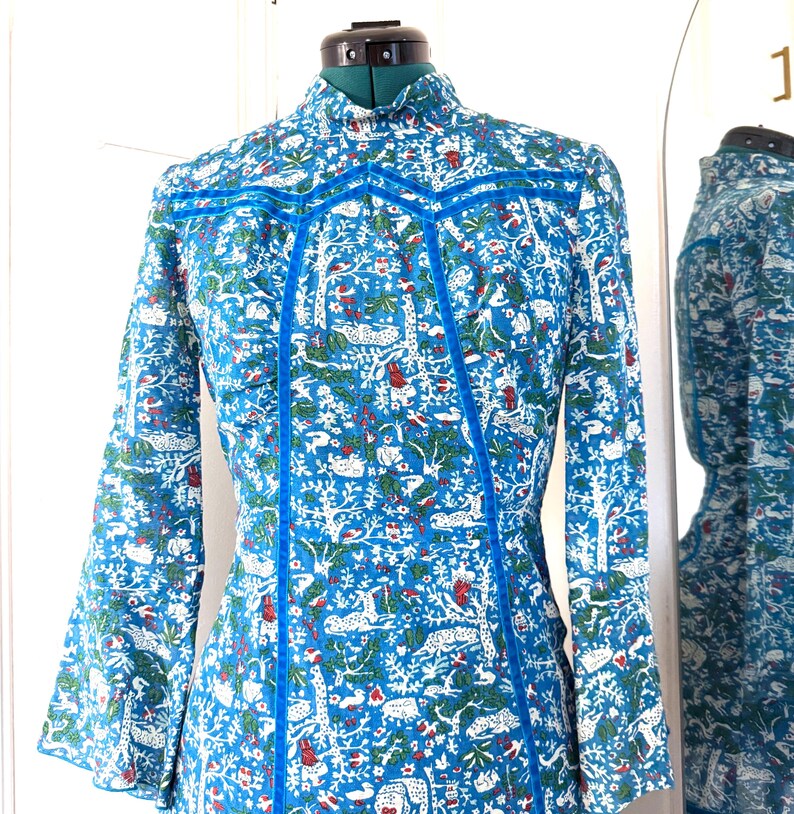 May include: A long-sleeved dress with a high collar, displayed on a mannequin. The dress features a vibrant blue background with a white, green, and red pattern of trees and animals. Bright blue trim accents the seams and neckline.