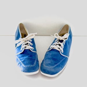 May include: A pair of blue canvas sneakers featuring a moccasin-style stitched toe, white laces, and a white rubber sole. The shoes have a low-top silhouette with light-colored interior lining and silver-toned eyelets for the lace-up closure.