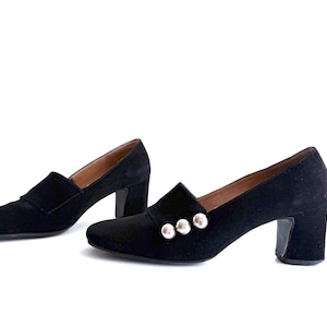 Op de afbeelding: Een paar zwarte su&egrave;de loafers met hak. De schoenen hebben een vierkante neus, een blokhak en zilveren knoopdetails. De schoenen staan tegen een witte achtergrond.