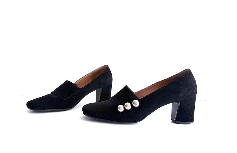 Op de afbeelding: Een paar zwarte su&egrave;de loafers met hak. De schoenen hebben een vierkante neus, een blokhak en zilveren knoopdetails. De schoenen staan tegen een witte achtergrond.