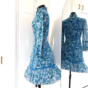 May include: A blue dress with a floral pattern and long sleeves. The dress has a high collar and a ruffled hem. The dress is displayed on a mannequin in front of a mirror, showing the back of the dress.