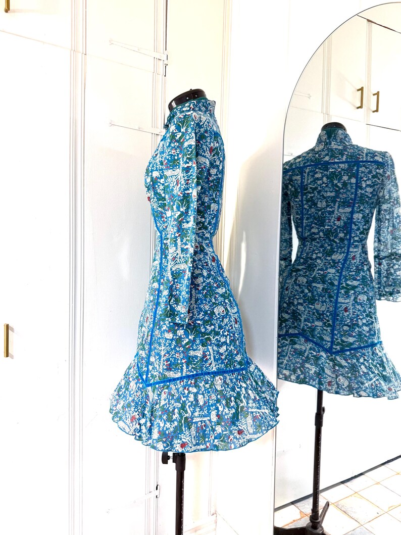 May include: A blue dress with a floral pattern and long sleeves. The dress has a high collar and a ruffled hem. The dress is displayed on a mannequin in front of a mirror, showing the back of the dress.