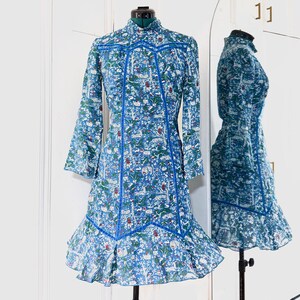 May include: A blue dress with a floral pattern and long sleeves is displayed on a mannequin. The dress features a high collar, blue trim, and a ruffled hem. The dress is reflected in a nearby mirror.