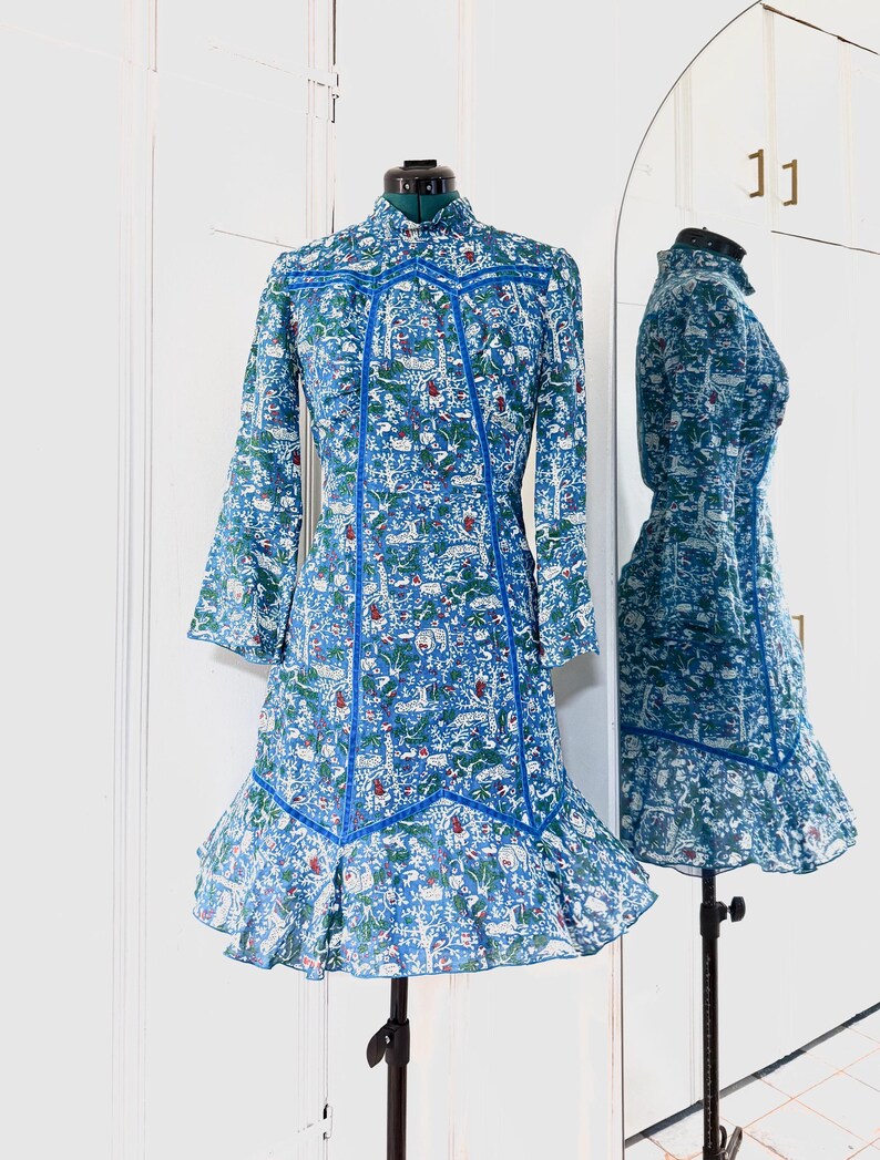 May include: A blue dress with a floral pattern and long sleeves is displayed on a mannequin. The dress features a high collar, blue trim, and a ruffled hem. The dress is reflected in a nearby mirror.