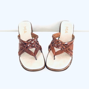 May include: A pair of brown leather sandals with a woven design. The sandals have a low heel and a thong strap. The brand name "MIA" is printed on the straps.