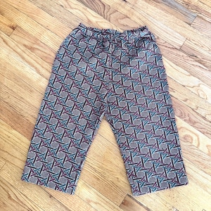 May include: A pair of patterned capri pants with an elastic waistband. The fabric features a geometric design in shades of teal, burgundy, and black. The pants are displayed on a wooden surface.