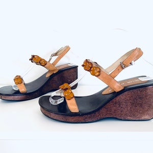 May include: A pair of brown leather wedge sandals with a buckle fastening and flower-shaped embellishments. The sandals have a wooden platform and a cork wedge heel.