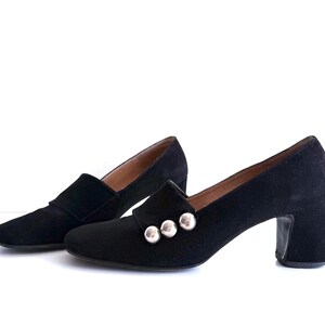 Op de afbeelding: Een paar zwarte su&egrave;de schoenen met hakken. De schoenen hebben een klassiek ontwerp met een ronde neus en een blokhak. Drie zilverkleurige knopen sieren de wreef van &eacute;&eacute;n schoen. De schoenen staan tegen een witte achtergrond.