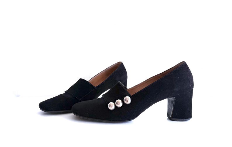 Op de afbeelding: Een paar zwarte su&egrave;de schoenen met hakken. De schoenen hebben een klassiek ontwerp met een ronde neus en een blokhak. Drie zilverkleurige knopen sieren de wreef van &eacute;&eacute;n schoen. De schoenen staan tegen een witte achtergrond.