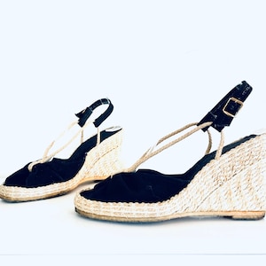 Op de afbeelding: Een paar zwarte suède sandalen met een wigvormige hak en touwdetails en een gespsluiting.