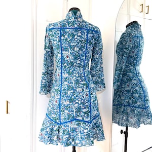 May include: A long-sleeved, blue dress with a whimsical print of animals and foliage. The dress features a high collar, blue trim, and a ruffled hem. The dress is displayed on a mannequin in front of a mirror.