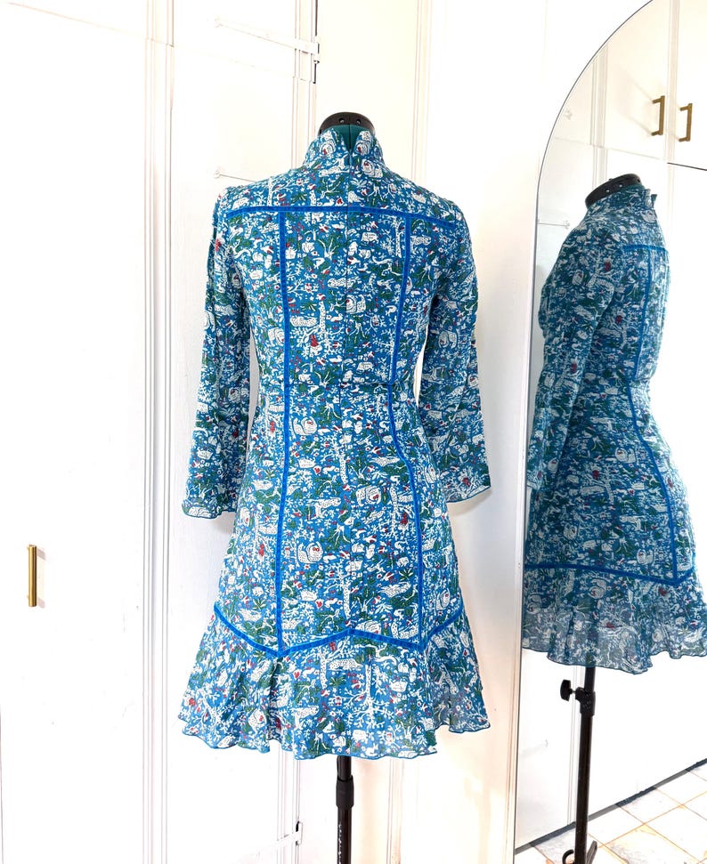 May include: A long-sleeved, blue dress with a whimsical print of animals and foliage. The dress features a high collar, blue trim, and a ruffled hem. The dress is displayed on a mannequin in front of a mirror.