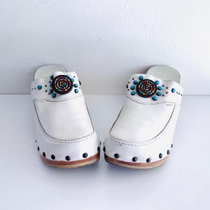 May include: A pair of white leather clogs with a decorative beaded design. The clogs feature a round, multi-colored beaded embellishment and turquoise beads. The soles are wooden with black studs around the edges.