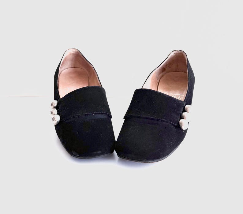 Op de afbeelding: Een paar zwarte su&egrave;de instappers. Elke schoen heeft een decoratief zilveren knoopdetail aan de zijkant. De schoenen hebben een lage hak en een ronde neus. De binnenkant van de schoenen is lichtbruin.