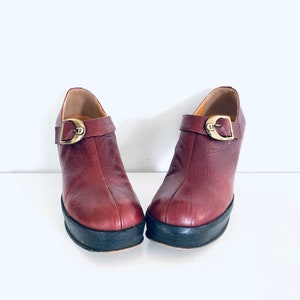Könnte beinhalten: Ein Paar dunkelrote Lederstiefeletten mit einer goldenen Schnalle an jedem Schuh. Die Stiefeletten haben eine Plateau-Sohle und einen dicken Absatz.