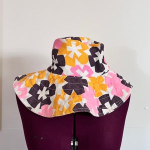 Op de afbeelding: Een brede zonnehoed met een witte achtergrond en een kleurrijk bloemenpatroon in roze, gele en bruine tinten.