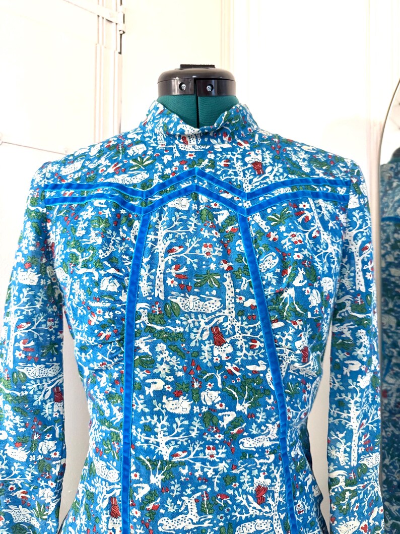 May include: A blue patterned dress with a high collar and long sleeves. The fabric features a white, red, and green forest design. The dress is accented with blue velvet trim.