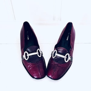 May include: A pair of burgundy leather loafers with silver buckle accents. The loafers have a pointed toe and a low heel.