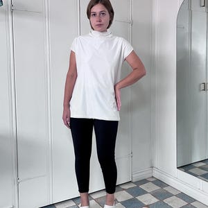 May include: A woman wearing a white mock neck shirt with short sleeves and black leggings. She is wearing white clogs and standing on a checkered floor.