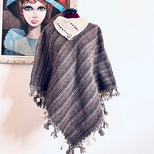 May include: A woven poncho in shades of brown, gray, and white, displayed on a mannequin. The poncho features a cream-colored collar with a wooden button and fringe detailing along the edges. A painting hangs in the background.