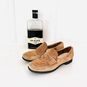 May include: A pair of brown suede loafers with a diamond-shaped design on the top. The loafers are on a white surface next to a bottle of Mr. Black Cold Brew Coffee.