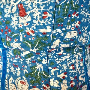 May include: Close-up of a blue fabric with a white, red, and green pattern. The pattern includes stylized trees, birds, and other natural elements. The fabric has a textured appearance and is trimmed with a blue ribbon.