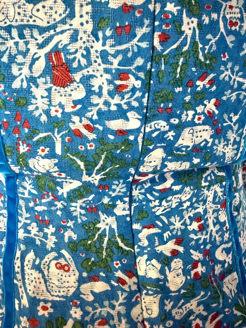May include: Close-up of a blue fabric with a white, red, and green pattern. The pattern includes stylized trees, birds, and other natural elements. The fabric has a textured appearance and is trimmed with a blue ribbon.