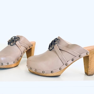 Puede incluir: Un par de zuecos de cuero gris con tacón, plataforma y tacón de madera. Los zuecos tienen una hebilla de metal decorativa en la correa.