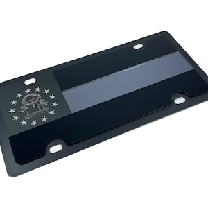 May include: Black metal license plate frame with a silver embossed design of the state of Georgia seal. The seal features a dome with three figures holding a banner that reads "Wisdom, Justice, Moderation". The seal is surrounded by 13 stars.