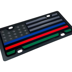 May include: Black license plate featuring a thin blue line American flag design. The flag is grey, blue, red, and green.